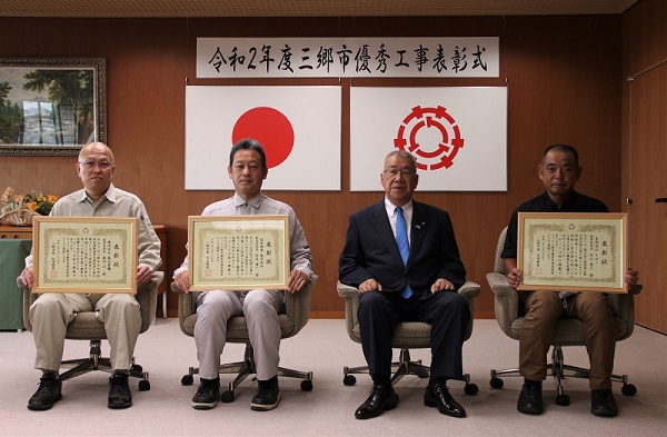 3名の男性が木津市長をはさんで椅子に座り、表彰状を持って記念撮影をする写真