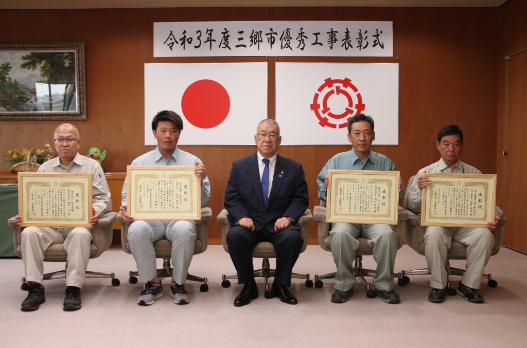 4名の男性が木津市長をはさんで椅子に座り、表彰状を持って記念撮影をしている写真