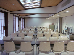 白い壁と障子風の窓、木目の天井の室内に、白い椅子と茶色の長机のセットが複数並べられた本館待合室の写真