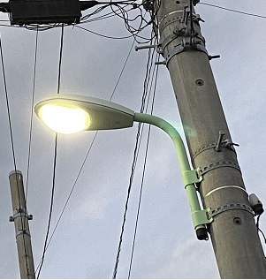 薄暗い曇り空の中光る電柱に設置された照明灯を下から写している写真