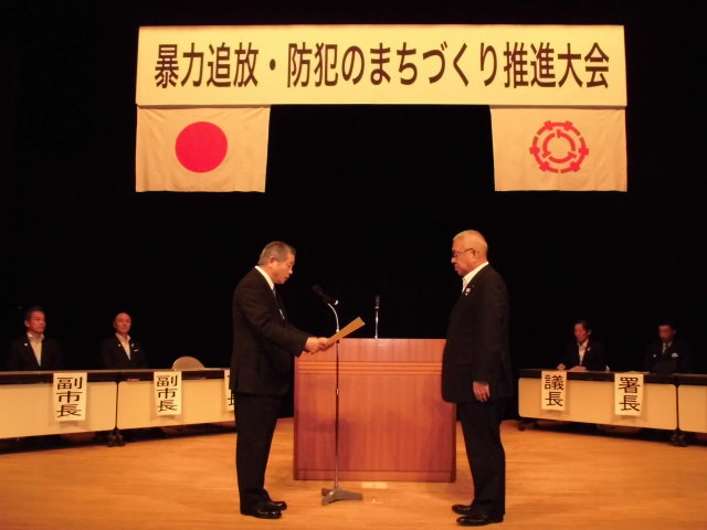 暴力追放・防犯のまちづくり推進大会と書かれた横断幕の下に日本国旗と三郷市章が飾られ、関係者が左右に分かれ並んで座るステージ上で、演壇の前に2名の男性が立ち、左側の男性が表彰状を読み上げ、右側の男性が聞いている様子の写真