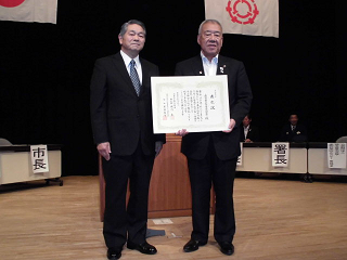 2名の男性が演壇の前に並び、受け取った表彰状を広げ撮影された記念写真