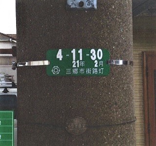 緑色のプレートに白色で管理番号や三郷市街路灯と書かれている電柱を撮影している写真