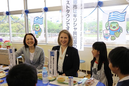 給食が用意された机で子ども達と向かい合ってにこやかに話をする駐日ギリシャ大使夫人の写真