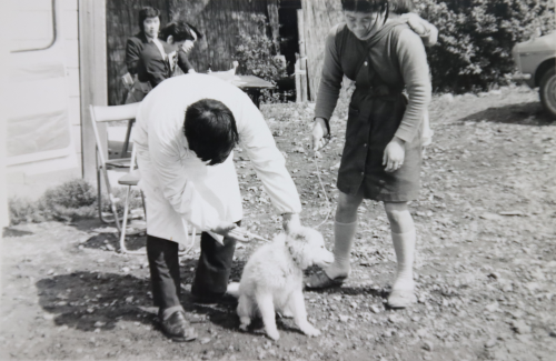 白衣を着用した男性がリードをつけた飼い犬の首輪を持ち予防接種を行っている様子を飼い主の女性が心配そうに見つめている白黒写真