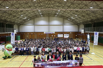 体育館で「ギリシャ文化交流プログラム」の横断幕が掲げられ、駐日ギリシャ大使夫人と木津市長を中心に関係者やかいちゃんのマスコット、多くの小学生たちが集合している記念写真