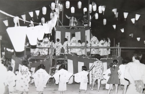 櫓の上で太鼓を叩いている音に合わせて浴衣姿の子どもや大人が手をつないで踊っている白黒写真