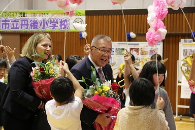 小学生たちがペーパーフラワーと棒で作った花のアーチを両側から支え、花束を持った木津市長と駐日ギリシャ大使夫人が中を通っていく様子の写真