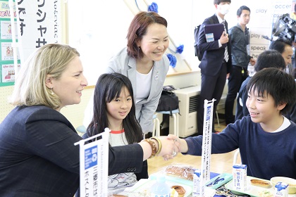給食が並んだ教室に関係者たちが集まる中、一人の男子生徒とにこやかに握手をしている駐日ギリシャ大使夫人の写真