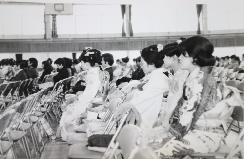 体育館内に並べられたパイプ椅子に座っている振り袖姿の女性たちを左から撮影した白黒写真