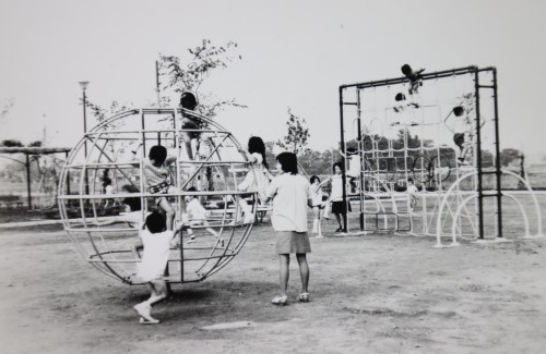 母親が見守るなか回転ジャングルジムなどの遊具で遊んでいる子ども達の白黒写真