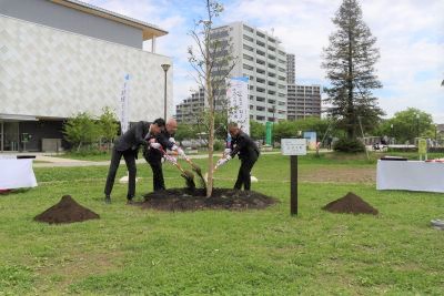 スーツを着た武居議長、木津市長、宅建協会 木村支部長が記念樹のシイノキにスコップで土をかけている様子の写真