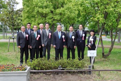 9人の式典参加者が横1列に並び、植樹したサツキを前に記念撮影をしている写真