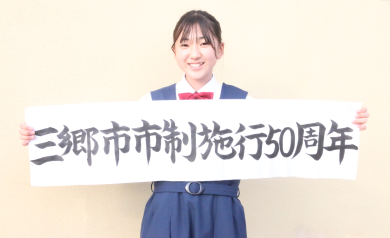 「三郷市市制施行50周年」の文字が筆で書かれた紙を両手で持っている題字制作者:江川 結衣さんの写真