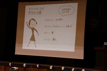 体育館に設置されたプロジェクタースクリーンに、4つの日本語がカタカナでギリシャ語の言葉に翻訳されモニターで映しだされている写真