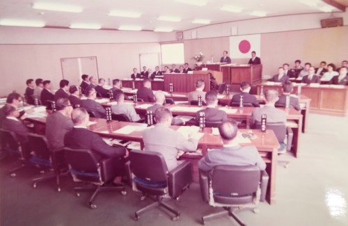 議場内に設置された檀上に男性が立ち、話をしているのを着席して聞いている議員の方々の後ろ姿の写真