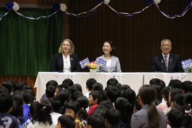 小学生たちが座って見守る中、体育館に設置された白いテーブルで、関係者の女性を間に挟み着席している駐日ギリシャ大使夫人と木津市長の写真