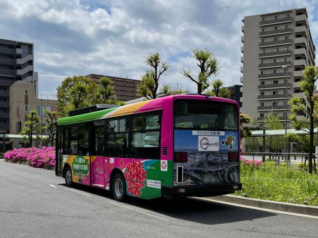 道路の脇に花壇があり、ピンクの花が咲いている横をラッピングバスが走っている様子の写真