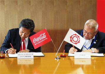 机にコカ・コーラ ボトラーズジャパン株式会社と三郷市市章のミニ旗が飾ってあり、関東営業本部長 井上豊理事(左)と木津市長(右)が協定書に署名をしている写真