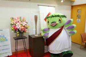赤い絨毯の中央に設置された台の上にオリンピックの聖火リレートーチが置かれ、横に立って記念撮影をしている古代ギリシャの衣装を着たかいちゃんの写真