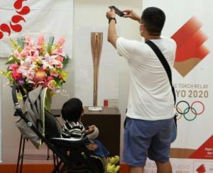 展示されている聖火リレートーチをスマートフォンで撮影している男性とベビーカーに乗った子どもを後ろから撮影した写真