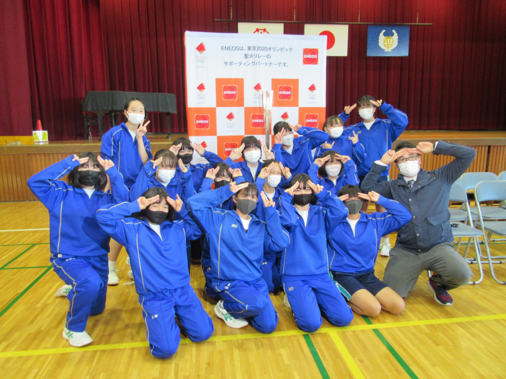青い学校ジャージを着たたくさんの女子学生と先生が展示されたトーチの前でピースサインをしている写真