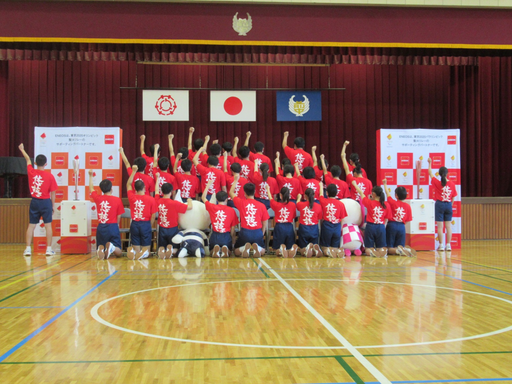 お揃いの赤いTシャツにショートパンツを履いたたくさんの学生たちが、体育館にある舞台の前で背中を向け左手に拳を作り突き上げているポーズをしている写真