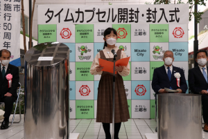 「タイムカプセル開封・封入式」と書かれた三郷市のバックボードの前で、タイムカプセルの横に代表の小学校4年生の女子児童が並んで作文を披露し、関係者の方々が注目している写真