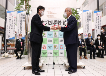 「タイムカプセル開封・封入式」と書かれた三郷市のバックボードの前で、タイムカプセルから取り出した手紙を市長から三郷郵便局長へ手渡している写真