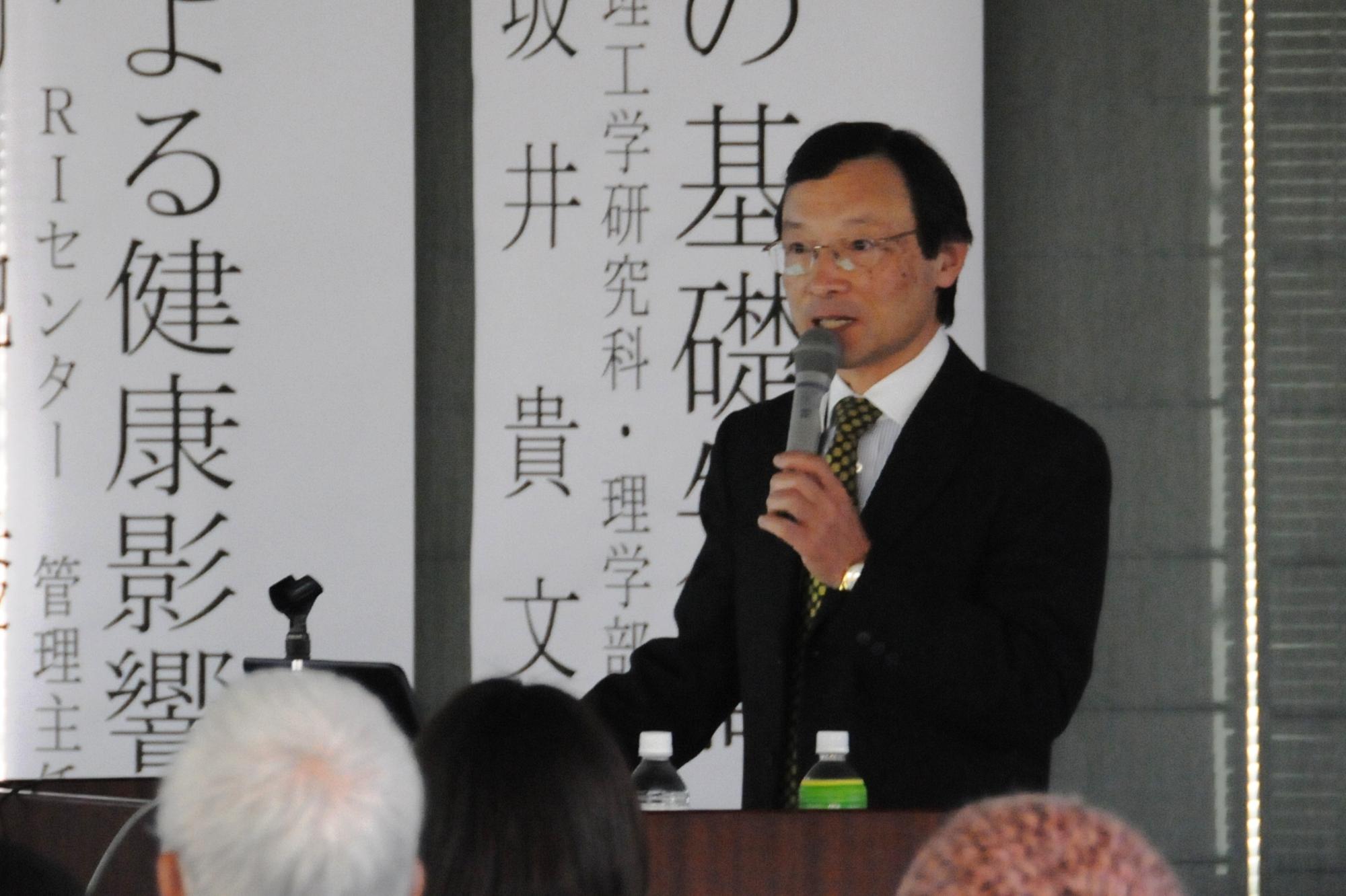 登壇に立ってマイクを持ち、放射線による健康影響について講演する菊池氏の写真