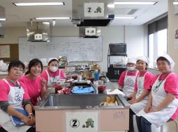 調理室の椅子に座りカメラ目線で微笑んでいる6人のエプロン姿の女性たちの写真