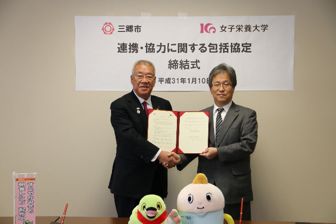 「三郷市 女子栄養大学 連携協力に関する包括協定締結式」と書かれた横断幕が後方に掲示され、三郷市長と香川女子栄養大学学長が並んで立ち、二人で協定書を持ち、握手をして写っている締結式の写真