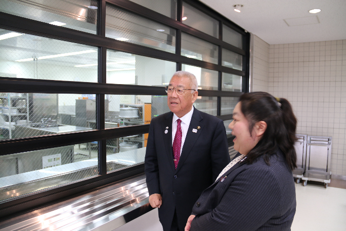関係者の女性と市長が学校内の調理室のような場所を窓越しに見学している写真