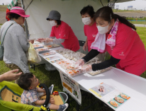 屋外に設置されたテント内で3名のヘルスメイトスタッフのかたが品物をイベント来場者に手渡そうとしている写真