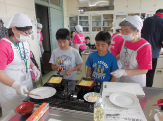 白のエプロン姿の女性たちが、男の子2人が卵焼きをフライパンで作っている様子を見守っている写真