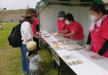 屋外に設置されたテント内でヘルスメイトスタッフのかたがイベント来場者の親子連れに品物を手渡そうとしている写真