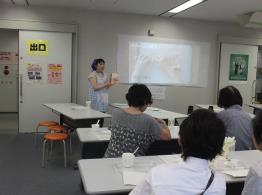 白の長テーブルが設置された会場の壁側で話をしている女性と、椅子に座って話を聞いている参加者たちの写真