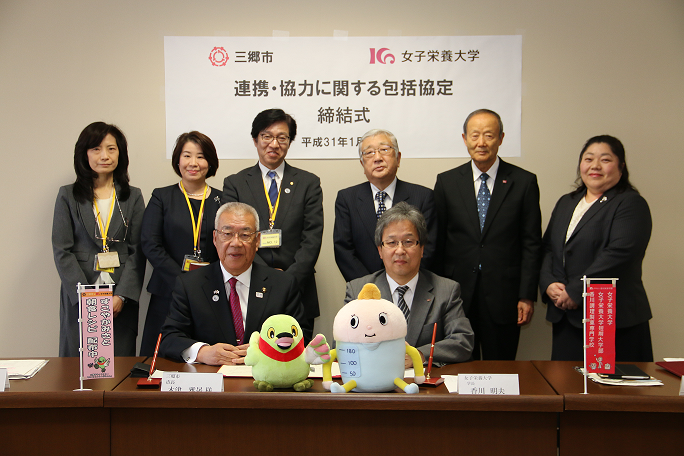 「三郷市 女子栄養大学 連携協力に関する包括協定締結式」と書かれた横断幕が後方に掲示され、三郷市長と香川女子栄養大学学長が並んで机に座っており、その後ろに6名の関係者の方々が並んで写っている集合写真