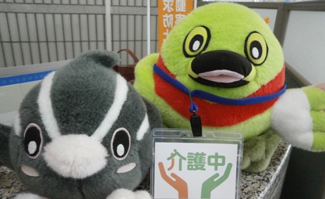 左につぶちゃん、右にかいちゃんのぬいぐるみが並んでおり、介護マークを入れた透明のケースをつけたネックストラップがかいちゃんの首に掛けられている写真