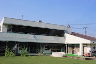 芝生の園庭の奥に、L字型で白い壁の、ベランダのある2階建ての建物の彦成保育所の外観写真