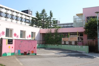 ピンク色の壁や外壁に桜の木や花のイラストが描かれている、道路に面した駐車場から見たさくら保育所の外観写真