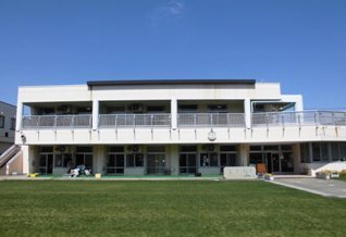 真っ青な空の下、芝生の園庭の奥にフェンスのついたベランダがある白っぽい壁をした2階建ての建物の早稲田保育所の外観写真