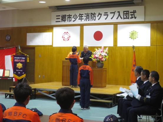 「三郷市少年消防クラブ入団式」の吊り看板が設置されている舞台上で、クラブ員の児童に消防手帳とクラブ員章を交付する市長の写真