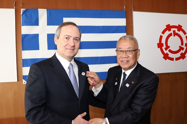 壁に掛かったギリシャ国旗の前で、フラワーホールに友好の証となるバッジを付けた駐日ギリシャ共和国大使と三郷市長が握手をしている記念写真