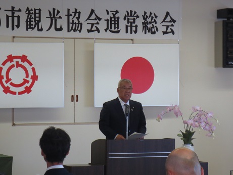 「三郷市観光協会通常総会」と書かれた横断幕と国旗と三郷市旗が飾られた室内に設置された演壇で挨拶を行っている市長の写真