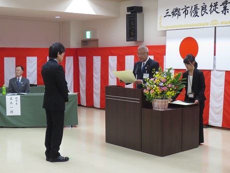 ピンク・黄色・紫の花々で彩られた花台と並ぶ演台に立ち表彰状を読み上げる三郷市長と横に立つ介添人、演台の前に立つ受賞者の男性の写真