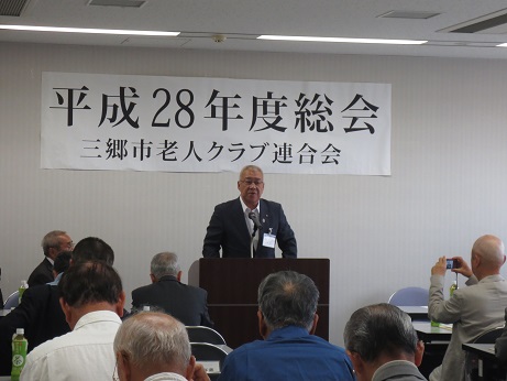 「平成28年度総会三郷市老人クラブ連合会」と書かれた横幕が壁に貼られ、長机が並んだ席に座っている参加者と向かい合って設置された演壇に立っている市長が挨拶を行っている写真