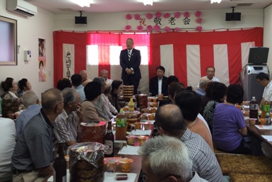 「祝敬老会」と書かれた戸ヶ崎一丁目下町会会館の紅白幕の前に立って挨拶を行っている市長の話を飲み物や料理が並んだテーブルの席に座って聞いている沢山の参加者の後ろ姿の写真