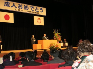 「成人おめでとう」」と書かれた横断幕や国旗と三郷市市旗が飾られたステージの檀上に立ち式辞を述べている市長の写真