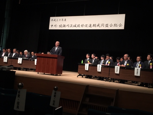 「平成30年度中川・綾瀬川流域改修促進期成同盟会総会」の吊り看板が設置されている舞台上に、多くの関係者たちが着席しており、市長が演壇に立って挨拶をしている写真